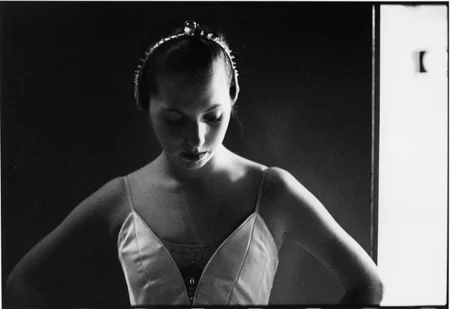 A young ballerina in a tiara looking down, hands resting on her hips