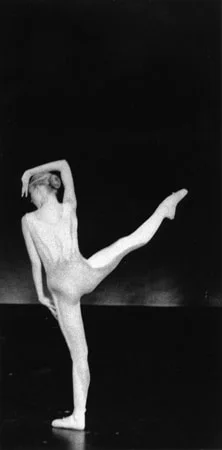 A dancer alone on a dark stage, one leg raised to the side and both arms curved overhead