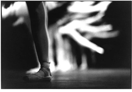 A dancer's foot in a ballet shoe held still while the skirts of other dancers blur behind