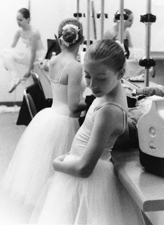 A young ballerina in a white tutu leaning on a counter backstage, other dancers behind her
