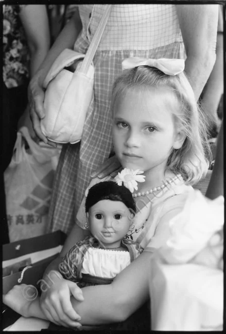 A young girl holding a doll on her lap, adults standing behind her