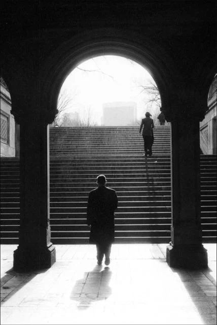 Two figures in silhouette beneath a stone arch, one climbing a broad flight of steps into the light