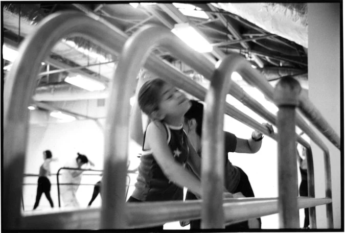 A young dancer stretching at the barre, seen through the bars of a second barre
