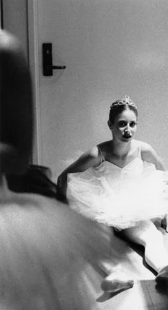 A ballerina in a tiara and tutu sitting on the floor of a backstage corridor, a figure blurred in the foreground