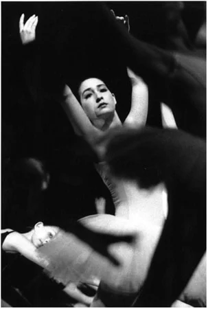 A dancer looking up with her arms raised, framed by the blurred limbs of the dancers around her
