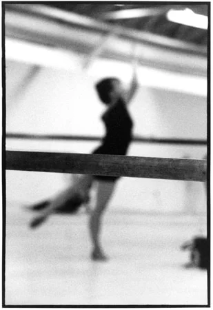 A dancer at the barre seen out of focus through a second barre in a bright studio