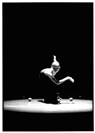 A dancer crouched at the centre of a lit circular stage, both arms reaching forward