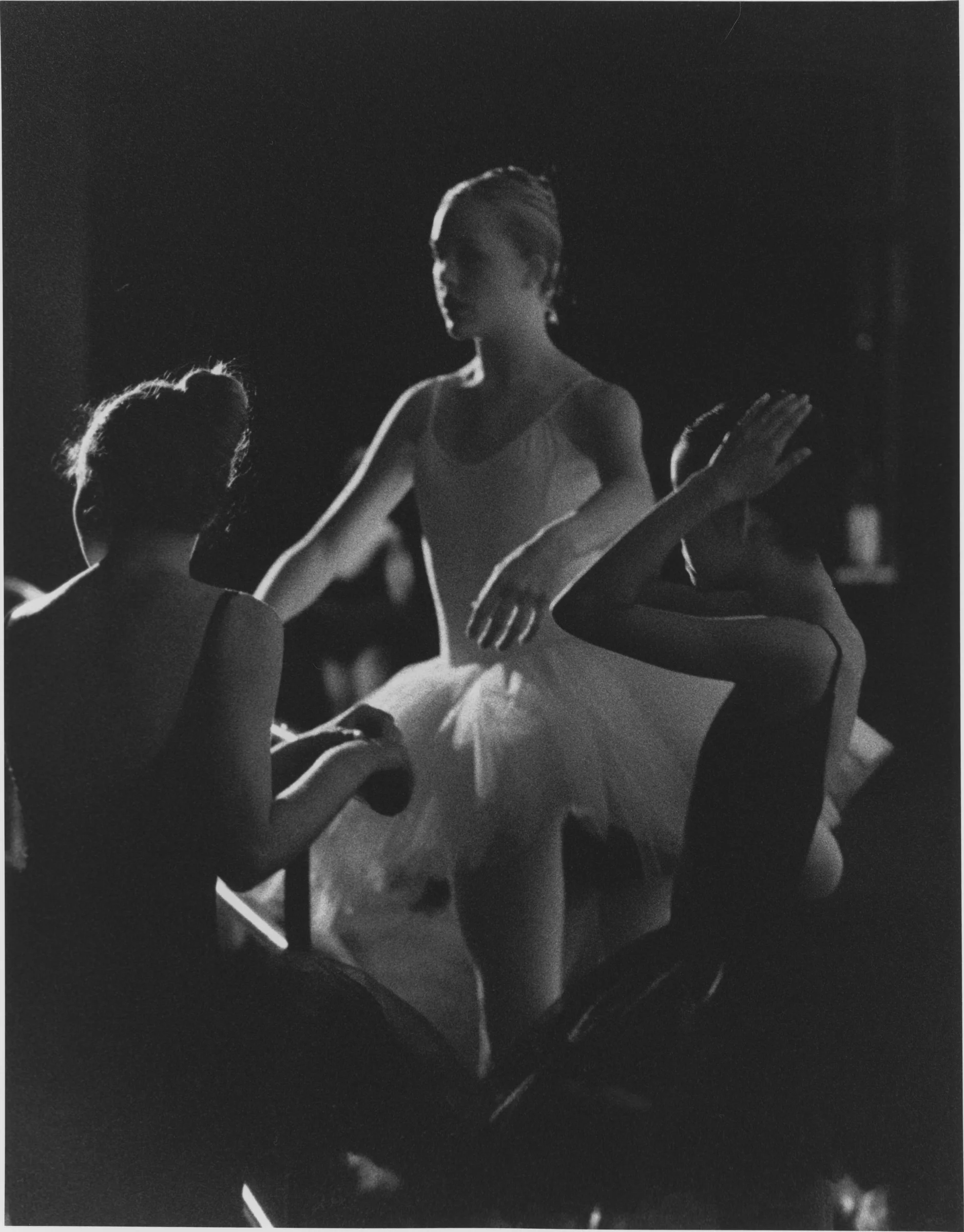 A ballerina standing among other dancers in the wings, her arms lifted