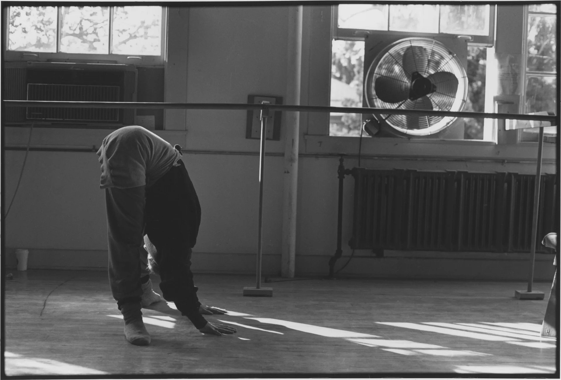 A figure bent to the floor of a sunlit rehearsal studio, barre and window fan behind