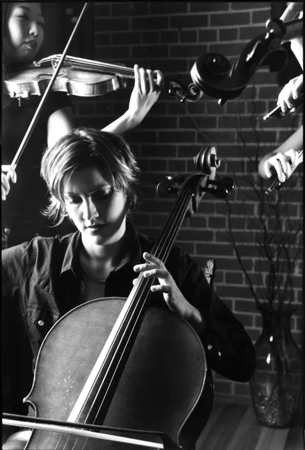 A cellist playing with her eyes lowered, a violinist behind her against a brick wall