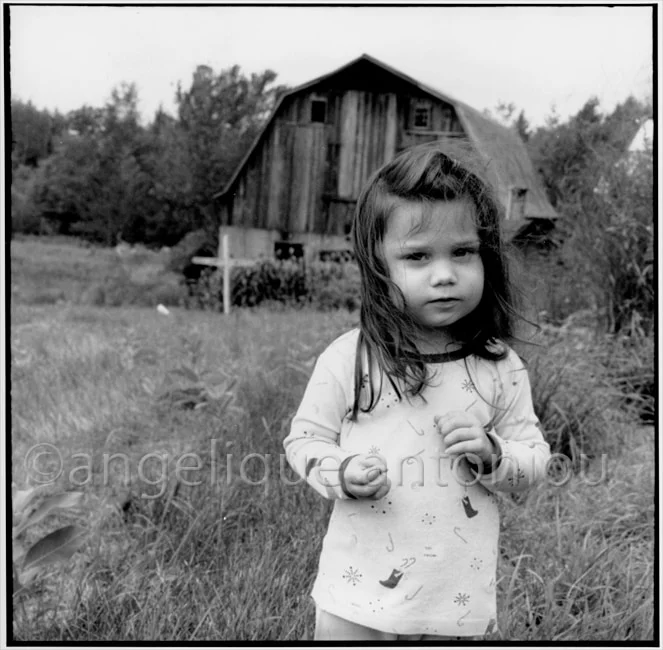 A young girl in a patterned nightdress standing in long grass before a weathered barn