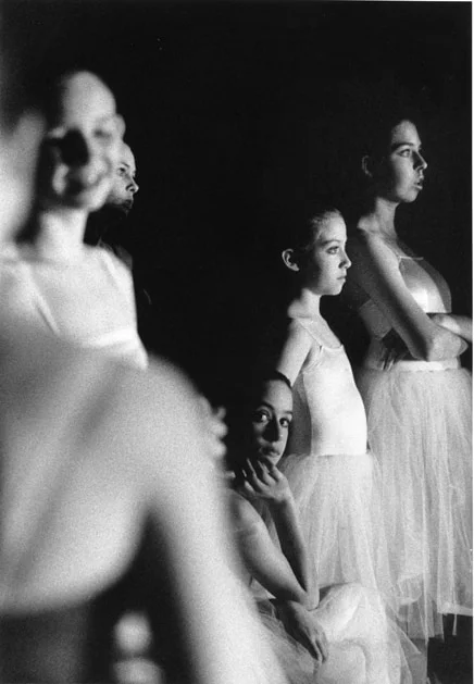 Ballerinas waiting in the wings, faces lit from the side, one resting her chin on her hand