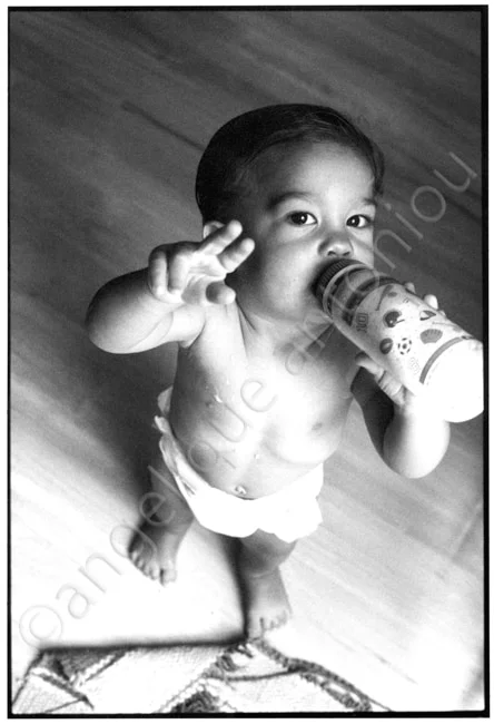 A baby standing on a wooden floor with a bottle raised, two fingers held up