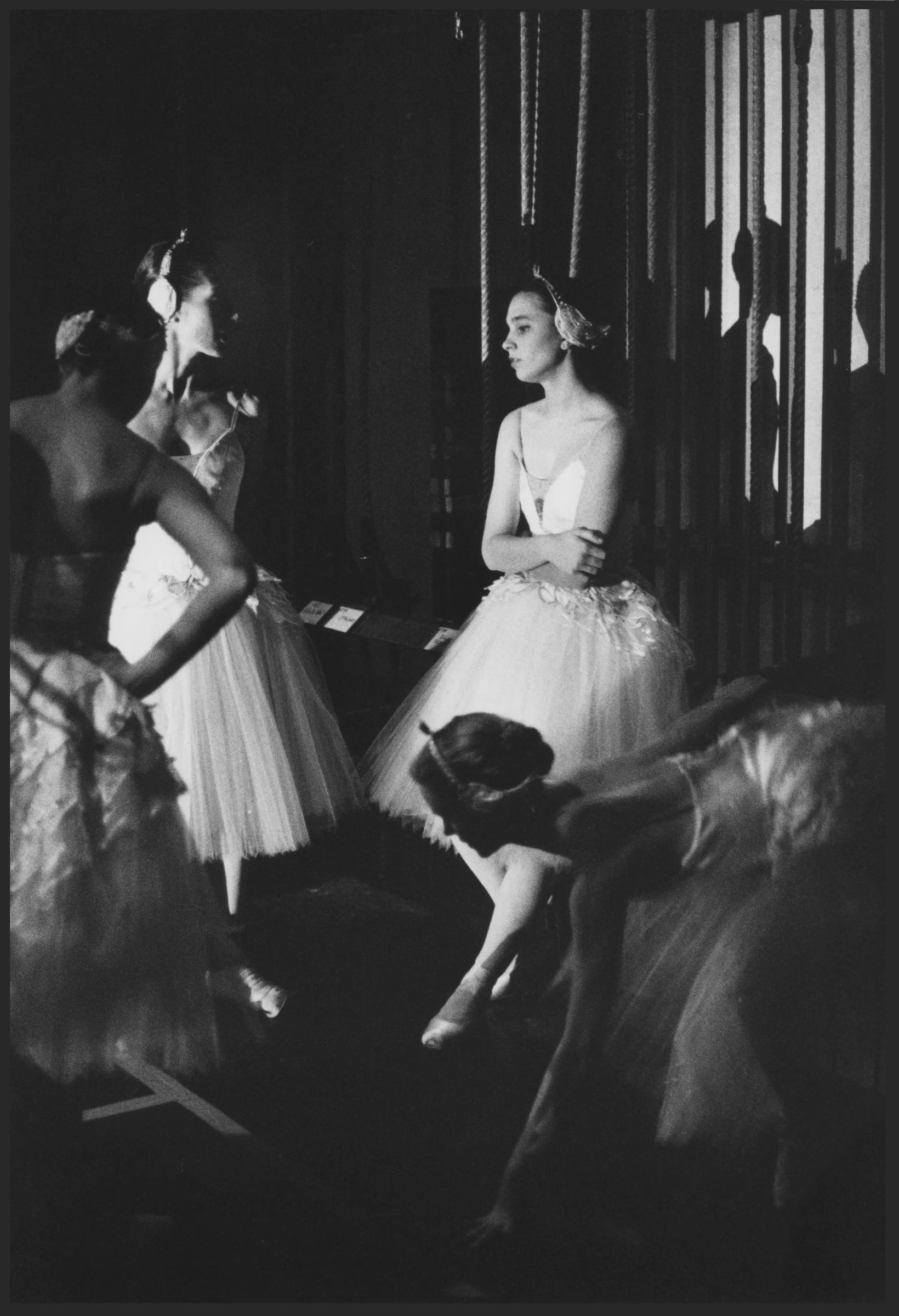 Ballerinas in long romantic tutus waiting in the wings, one bending to her shoe as another passes in a blur