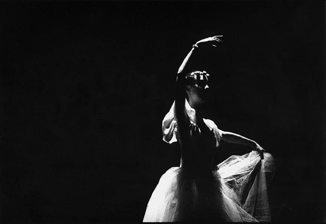 A ballerina lit from above in a pale tutu, one arm curved over her head against black