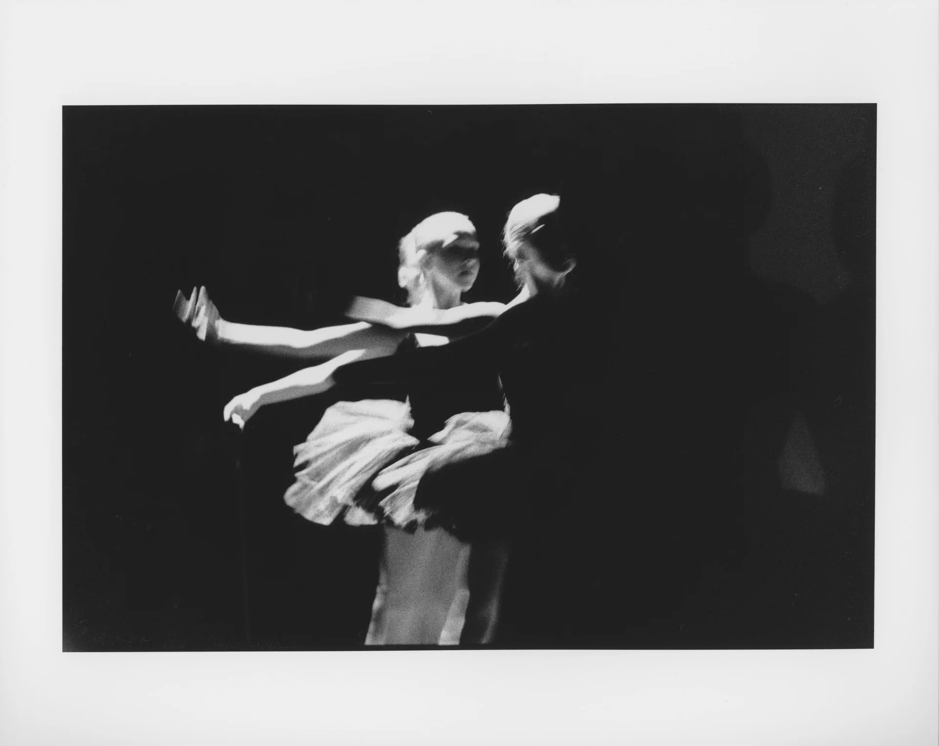 Two dancers together on a dark stage, blurred by the length of the exposure. Harsh downward light and shadows. High contrast ballet photograph. Ballet dancers on stage, arms in second position. Candid ballet photograph.