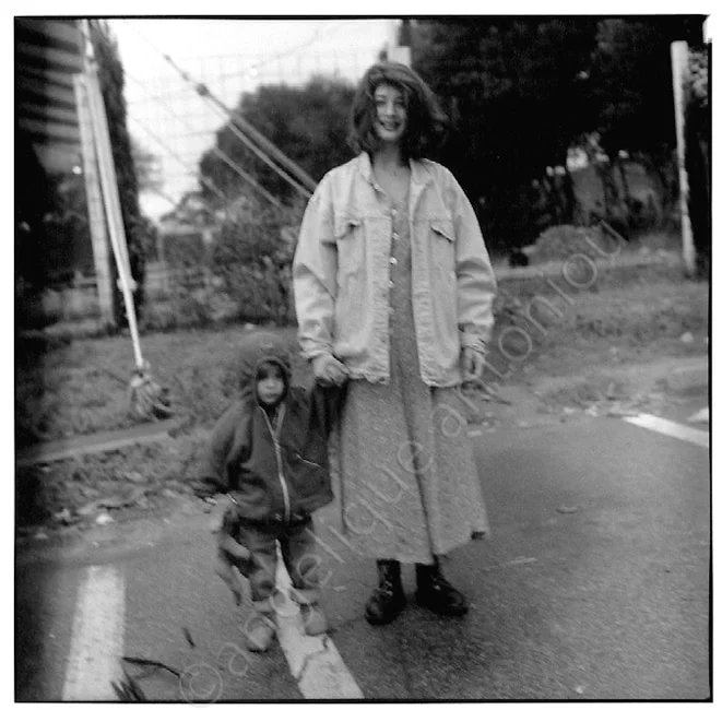 A young woman in a denim jacket holding a small child's hand at the edge of a road