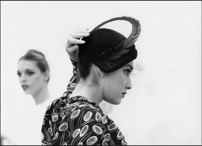 A woman in a feathered hat and patterned dress seen in profile, a second woman out of focus behind her