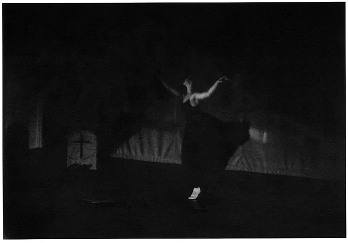 A dancer in a dark dress on a dimly lit stage, a gravestone flat standing behind her