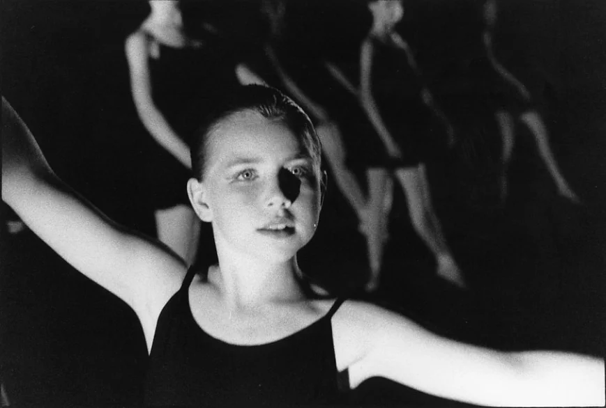 A young dancer with both arms outstretched lit from the front, other dancers moving in shadow behind