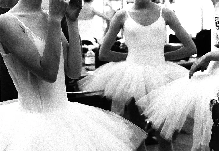 Dancers in white tutus crowded together in a dressing room, one raising a hand to her face