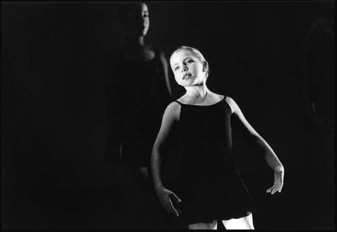 A young dancer in a dark shift lit from the front, another dancer shadowed behind her