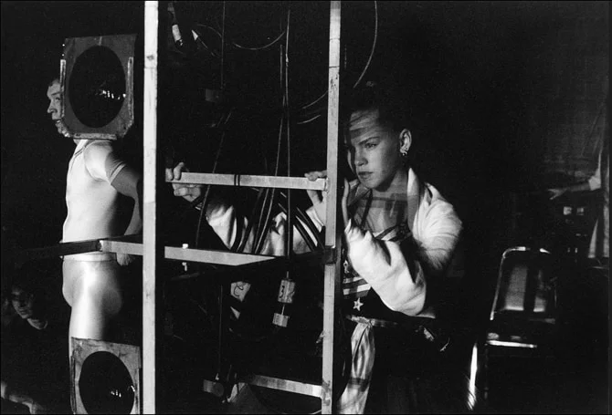 Dancers waiting behind a lighting scaffold and stage speakers in the wings