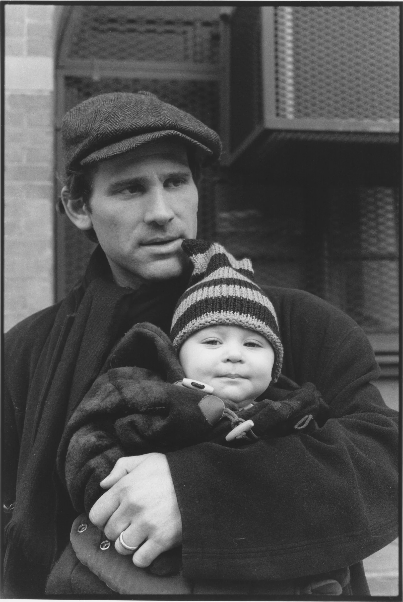 A man in a flat cap carrying a baby in a striped bobble hat against his chest on a city street. Reminiscent of a 1940s black-and-white photo.