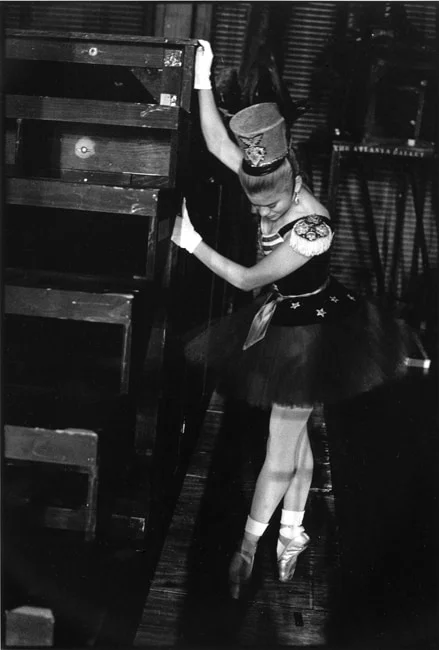 A ballerina in a military-style costume and tall hat rising to pointe beside stacked scenery flats