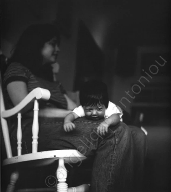 A baby leaning over an adult's knee, a woman in a rocking chair behind