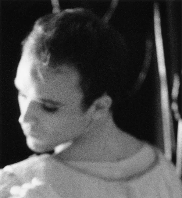 A male dancer in costume seen close in profile, looking down, backstage