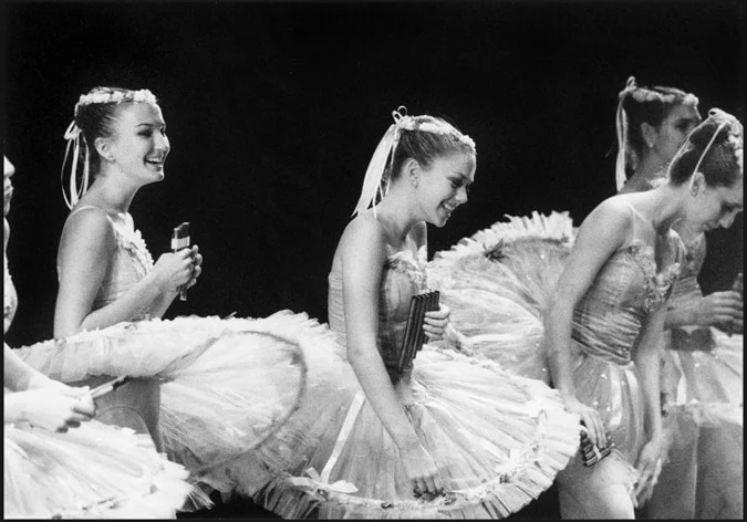 A line of ballerinas in tutus laughing together in the wings, one holding a lipstick