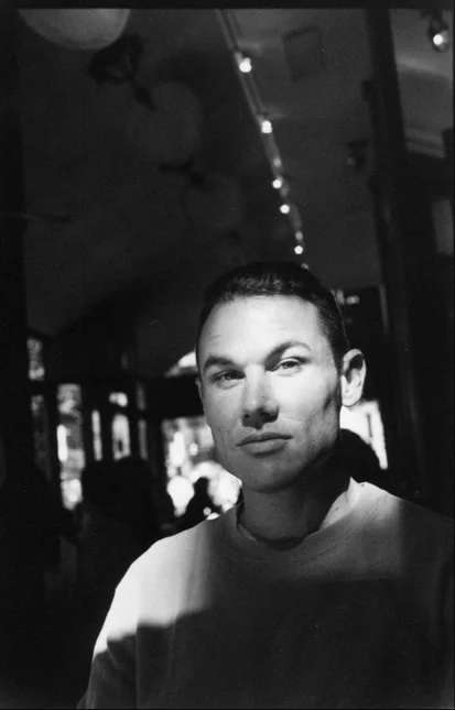 A man in a pale sweatshirt looking to camera in a cafe hung with paper lanterns