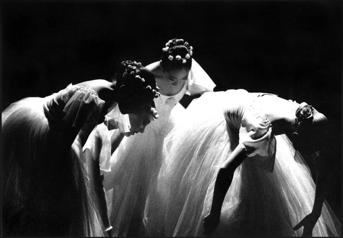 Three ballerinas in flowered headpieces bowing together into a pool of light on a dark stage