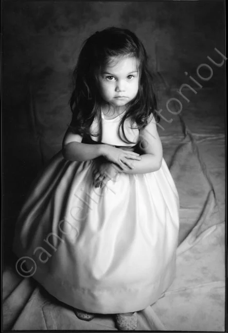 A small girl in a white dress sitting with her hands folded, looking straight to camera