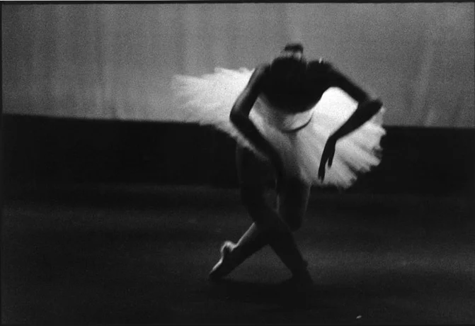 A ballerina bent low on a bare stage, arms crossed above her head