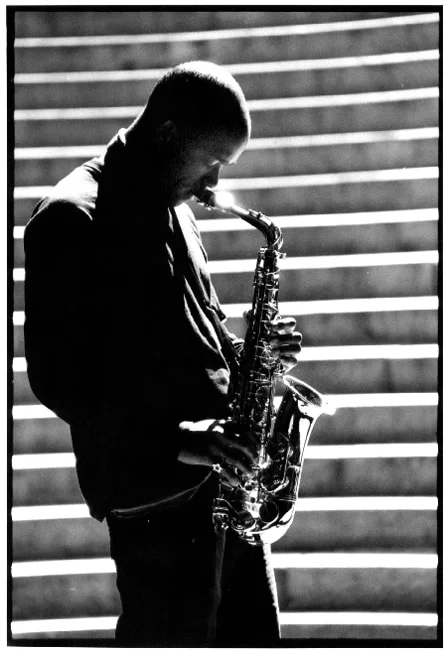 A saxophonist playing in profile against sunlit stone steps