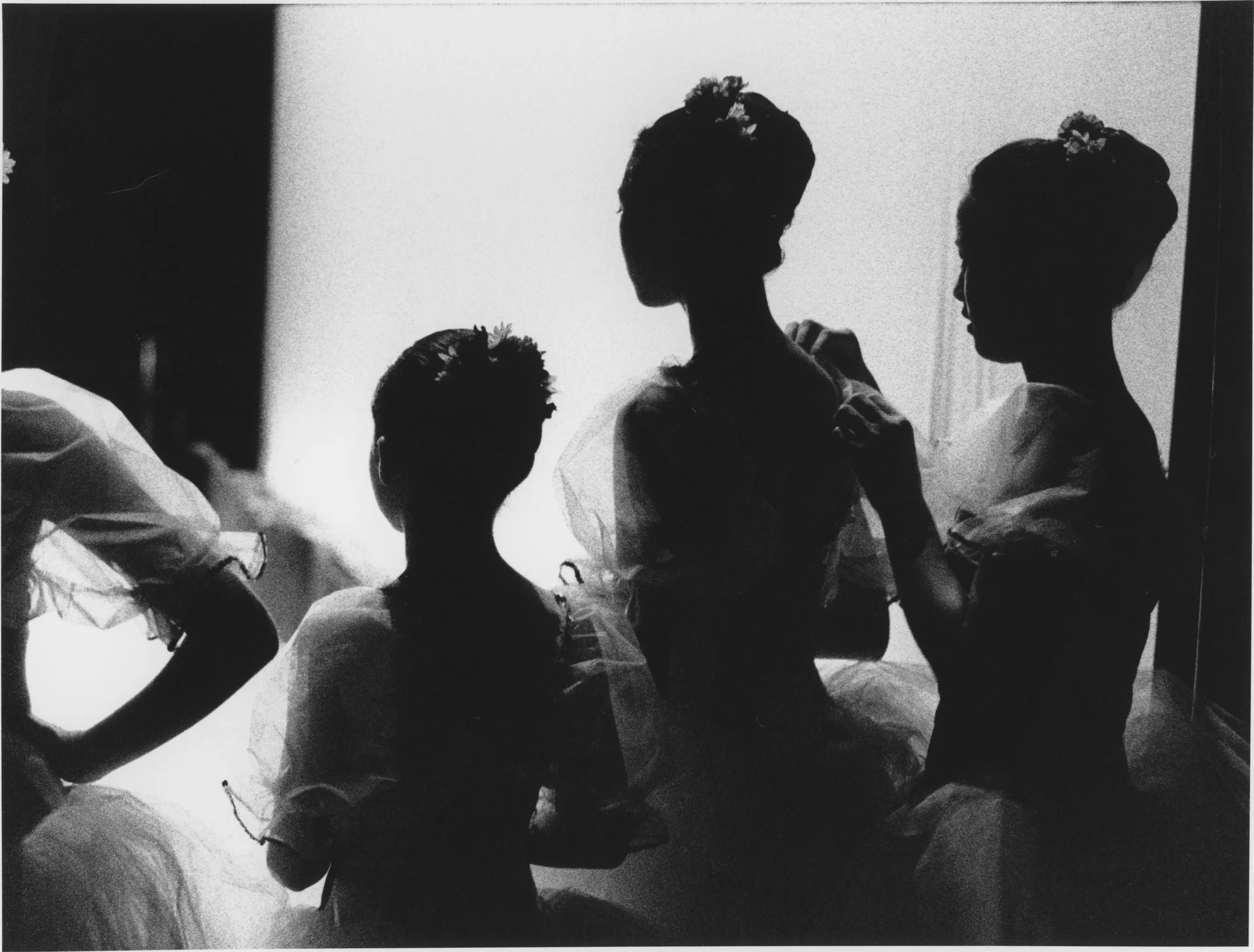 Ballerinas silhouetted against a bright wall, one reaching to fasten another's costume