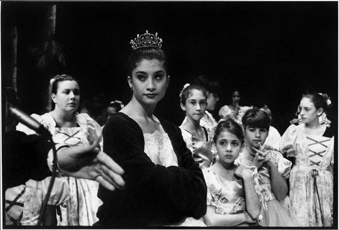 A young ballerina in a tiara and dark robe looking to camera, costumed children gathered behind her