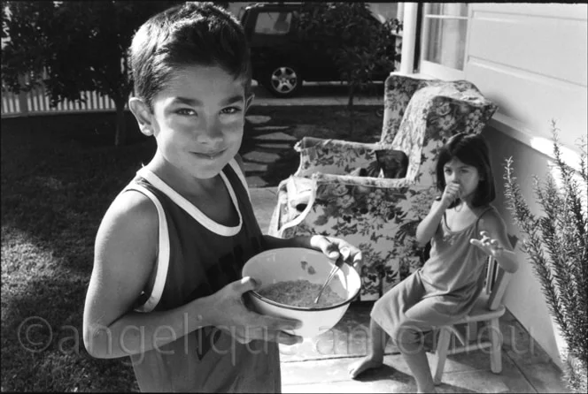 A boy on a porch holding a bowl of cereal, a younger girl eating on a chair behind him