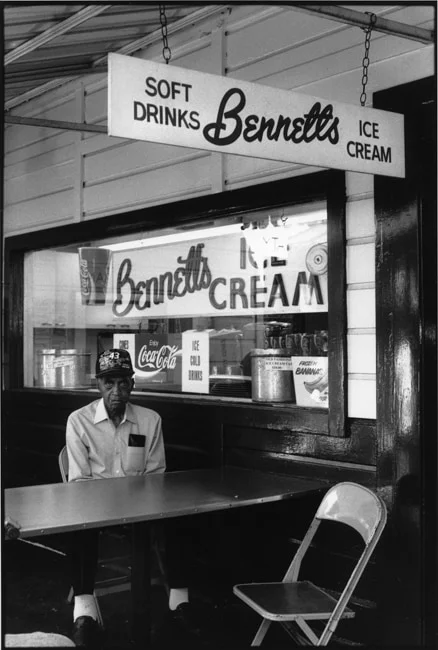 An elderly man sitting alone at a table outside a Bennett's ice cream counter, an empty folding chair opposite him