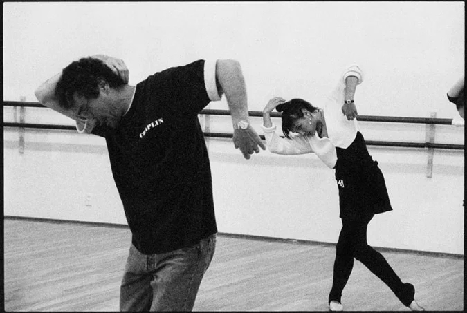 A teacher and a young dancer mirroring the same step, both bent sideways in a studio