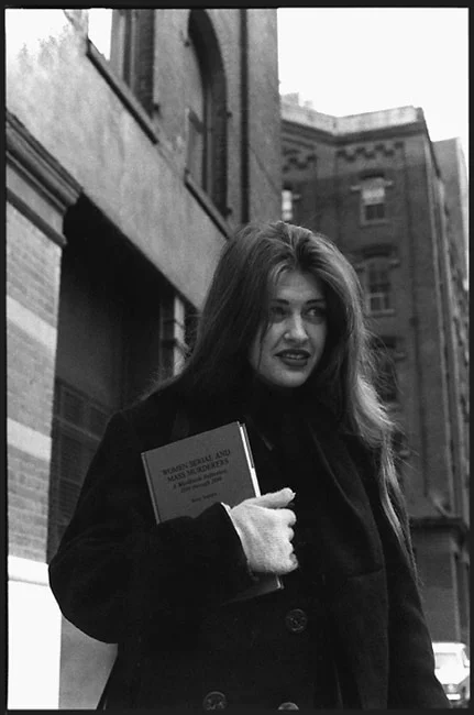 A young woman with long hair in a dark coat on a city street, holding a book against her chest