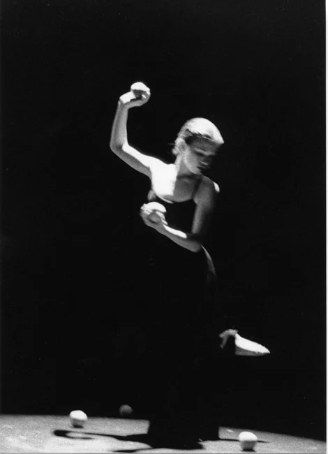 A dancer in a dark dress on a lit circular stage holding pale spheres, more scattered at her feet