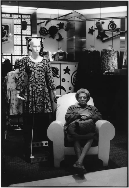 An older woman asleep in a white armchair in a clothing shop, a mannequin standing beside her