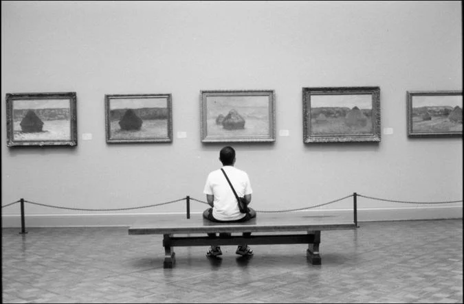 A single visitor sitting on a bench in a gallery facing a row of Monet haystack paintings