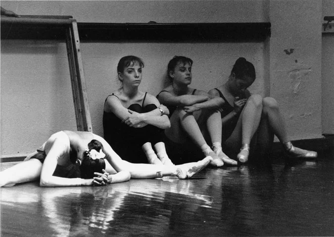 Four young dancers sitting against a studio wall in leotards while a fifth lies stretched out on the floor