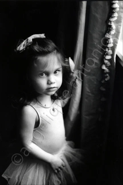 A small girl in a tutu with a bow in her hair standing beside a curtain